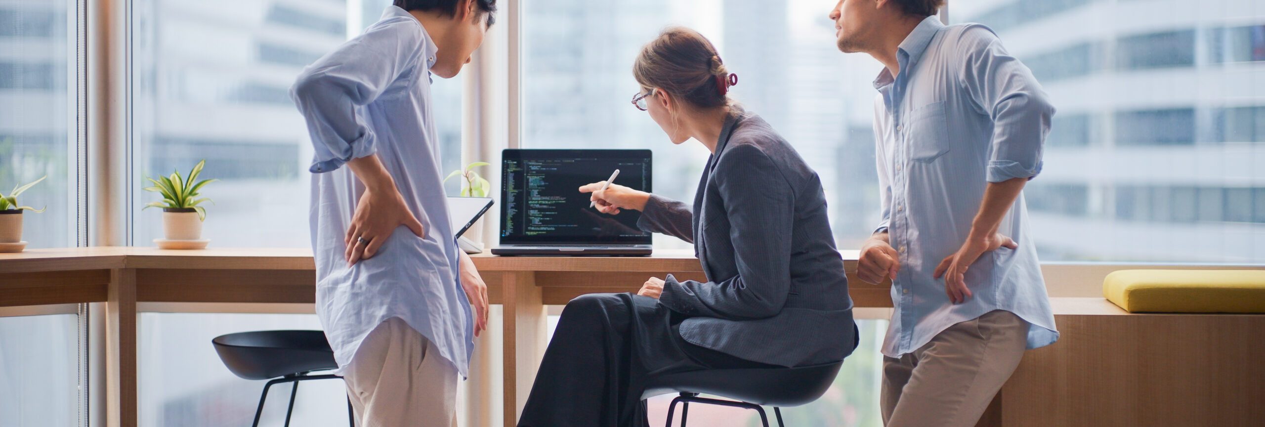 Three software engineers, including two men and one woman, are gathered around a laptop discussing programming code in a modern, high-rise office with large windows. The team appears to be reviewing or debugging software together, highlighting teamwork, collaboration, and the creative process in a tech startup or IT company. The cityscape outside the windows suggests a professional urban environment. This image conveys concepts of software development, agile workflow, modern business, innovation, and corporate teamwork.