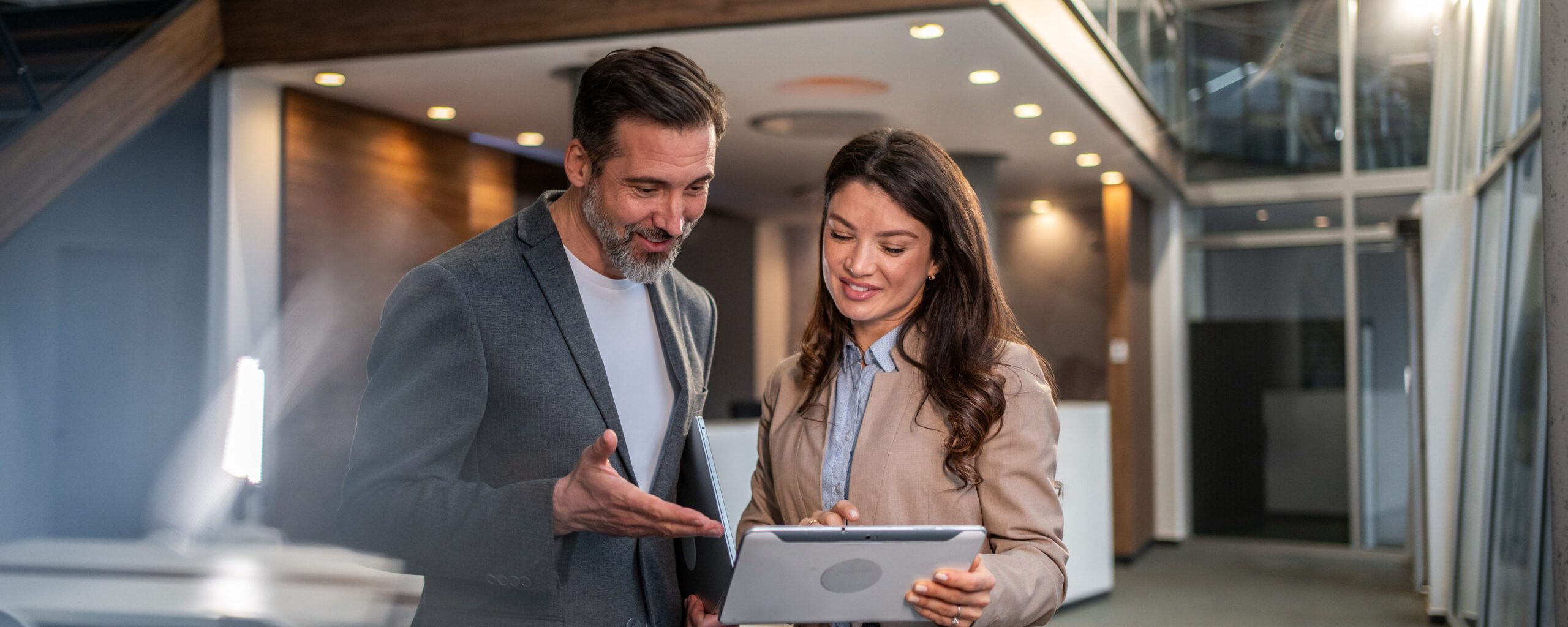 Two business professionals are smiling and engaging in discussion while reviewing content on a digital tablet, fostering teamwork and demonstrating modern technology use in a corporate environment
