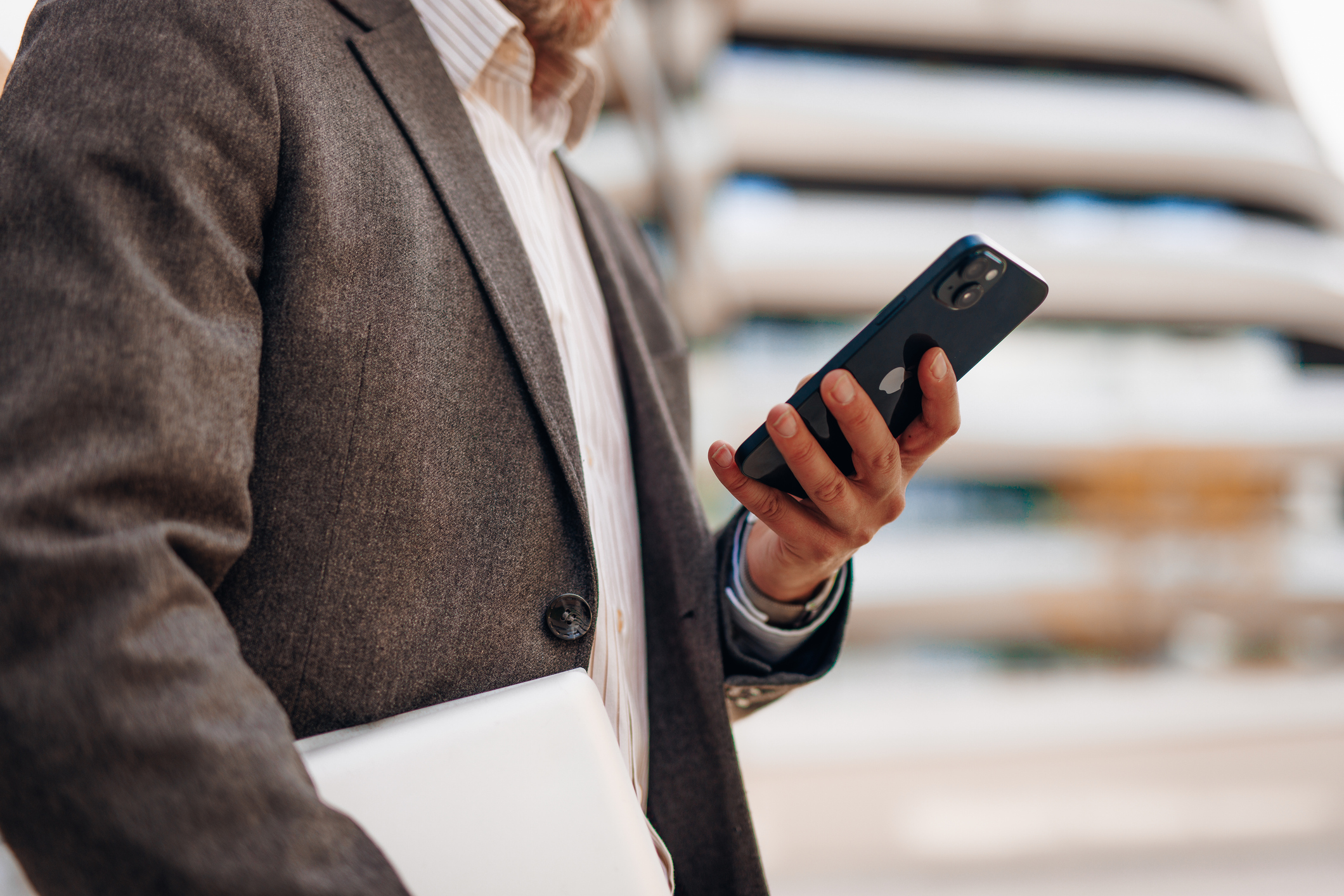 Businessman using smartphone and holding laptop in urban setting